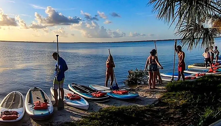 A group of people are preparing for paddleboarding near the water's edge at sunset.