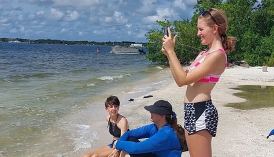 A person is standing and taking a selfie on the beach while two others sit by the water's edge.