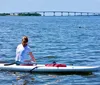 A person sits on a stand-up paddleboard on calm water with a bridge in the distance