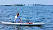 A person sits on a stand-up paddleboard on calm water with a bridge in the distance.