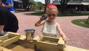 A young girl wearing sunglasses and holding a large amethyst geode smiles while standing at a gemstone mining sluice at an outdoor location.