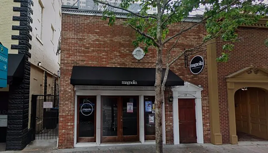 The image shows a storefront with a black awning labeled magnolia, a sidewalk tree in front, and adjacent buildings on a calm street.