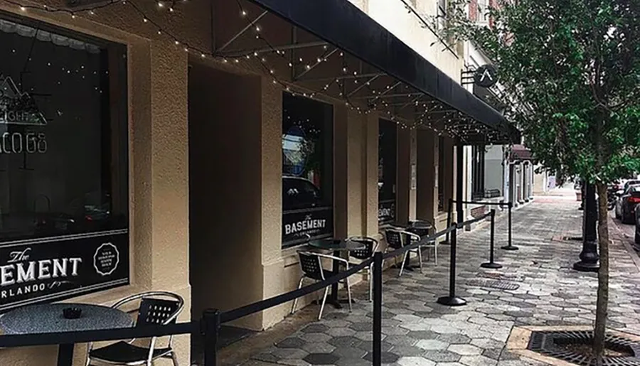 A quiet sidewalk cafe setting with string lights under an awning in an urban environment.