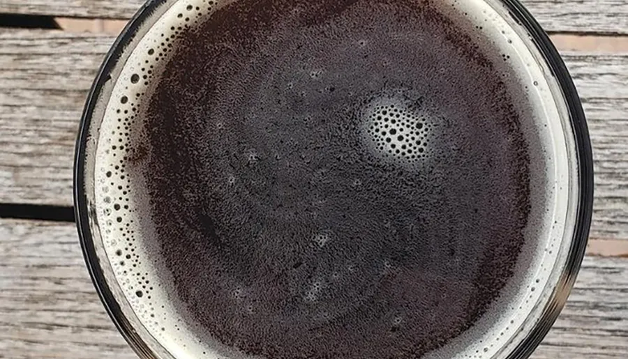 The image shows a top-down view of a dark beverage with a foamy head, possibly a stout or porter beer, in a glass placed on a wooden surface.