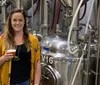 A woman is posing with a glass of beer in front of stainless steel brewing tanks in a brewery