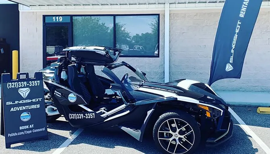 A black Polaris Slingshot three-wheeled vehicle is on display outside a rental establishment with advertising banners and a sign promoting adventures and bookings.