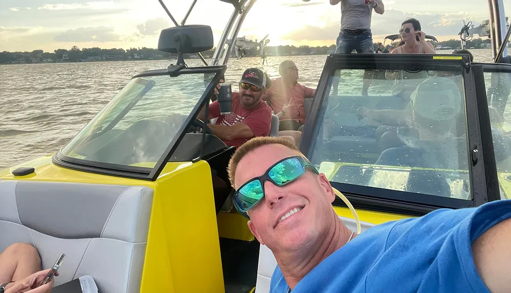 A group of people is enjoying a boat ride on a sunny day with some sitting at the back and one person taking a selfie in the foreground