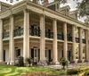 The image shows a large historic plantation-style house with a grand double-tiered columned front porch set among lush greenery and flowering plants
