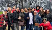 A group of smiling people pose together in front of a mural with artistic depictions of historical figures.