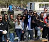 A group of happy people are posing for a photo together at a waterfront with a tour boat and some trees visible in the background