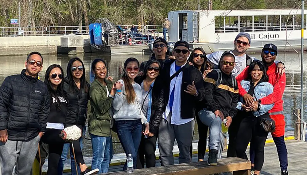 A group of happy people are posing for a photo together at a waterfront with a tour boat and some trees visible in the background