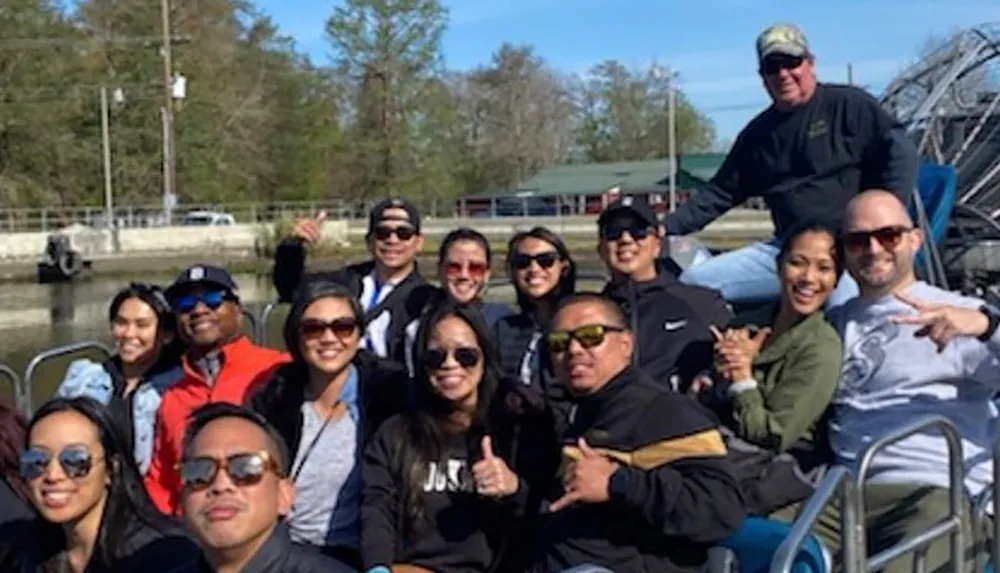 A group of cheerful people is posing for a photo together on a boat with some giving thumbs-up or peace signs appearing to be enjoying an outdoor excursion