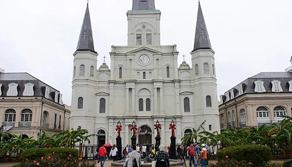 This image shows a group of people gathered in front of a large white cathedral with two spires and a clock flanked by classical buildings under an overcast sky