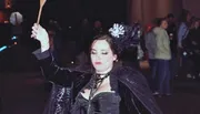 A person dressed in a dark, gothic-inspired costume with a wand is striking a dramatic pose during a nighttime event.