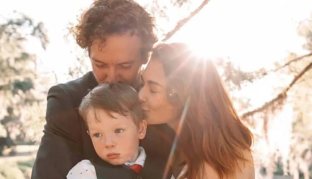 A family moment is captured with a man and a woman affectionately kissing a young child on the head in a sunlit outdoor setting