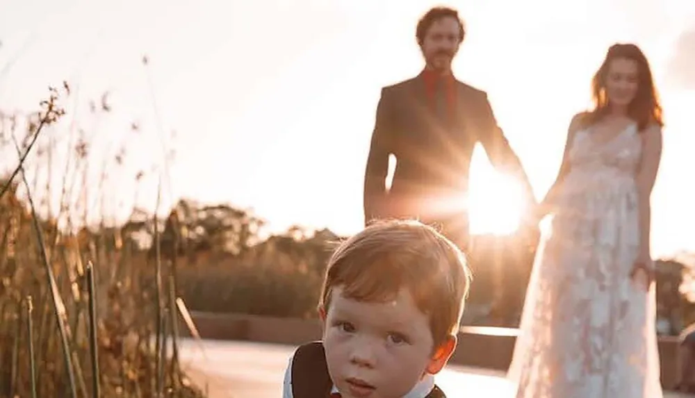 A young child in formal attire is in the foreground with a man and a pregnant woman holding hands in the background backlit by a warm sunset