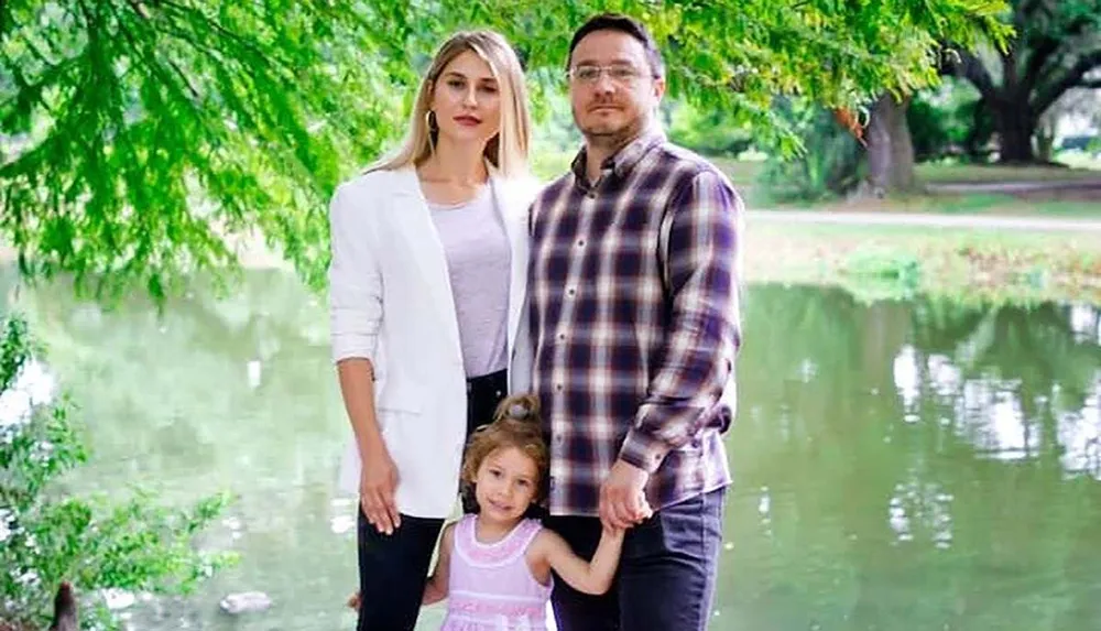 Two adults and a child pose together in a serene park with a pond and greenery in the background