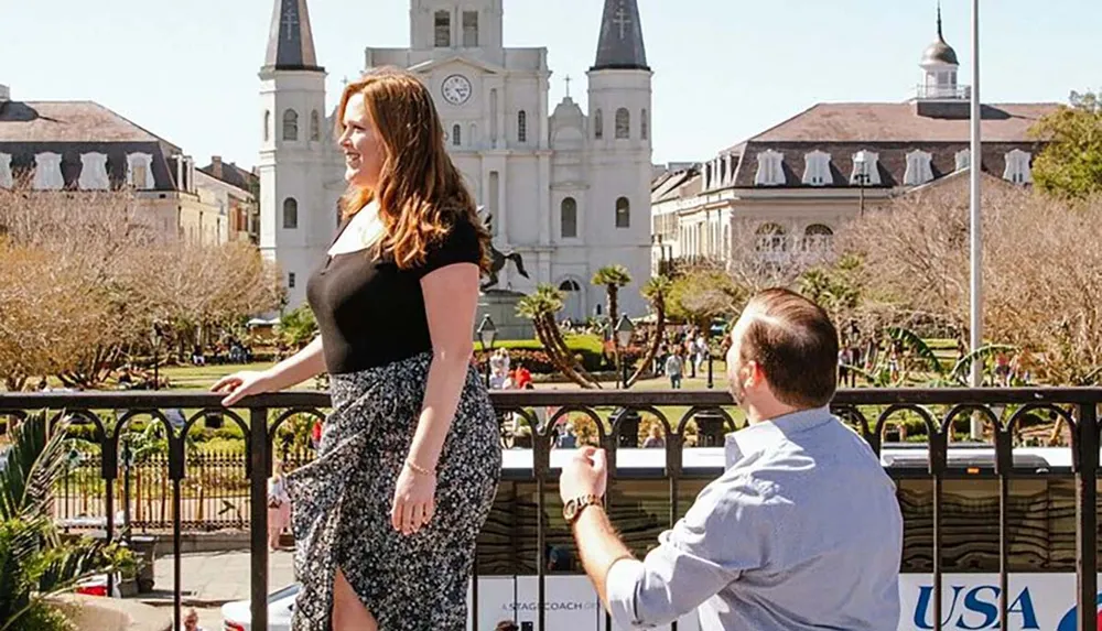 A man is kneeling in front of a woman in a picturesque outdoor setting possibly proposing with a historic building visible in the background