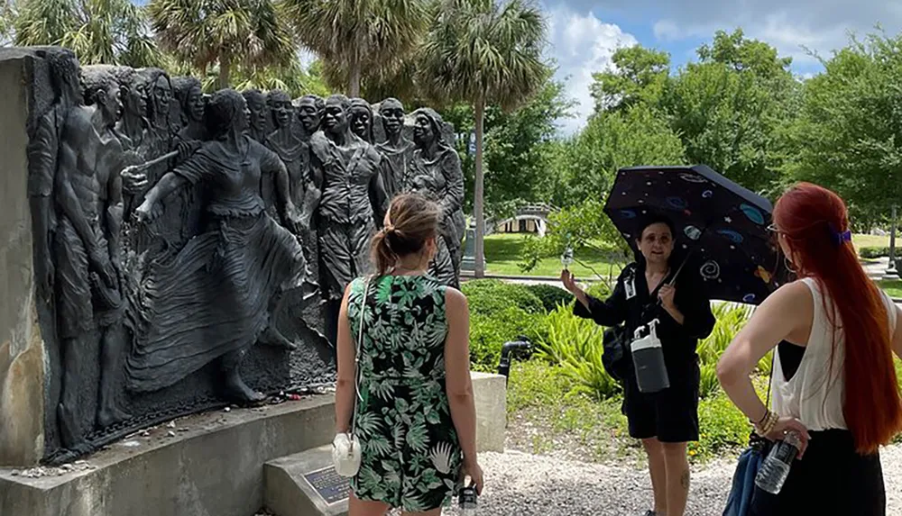 A group of tourists is listening to a guide with an umbrella who is explaining a relief sculpture in an outdoor setting