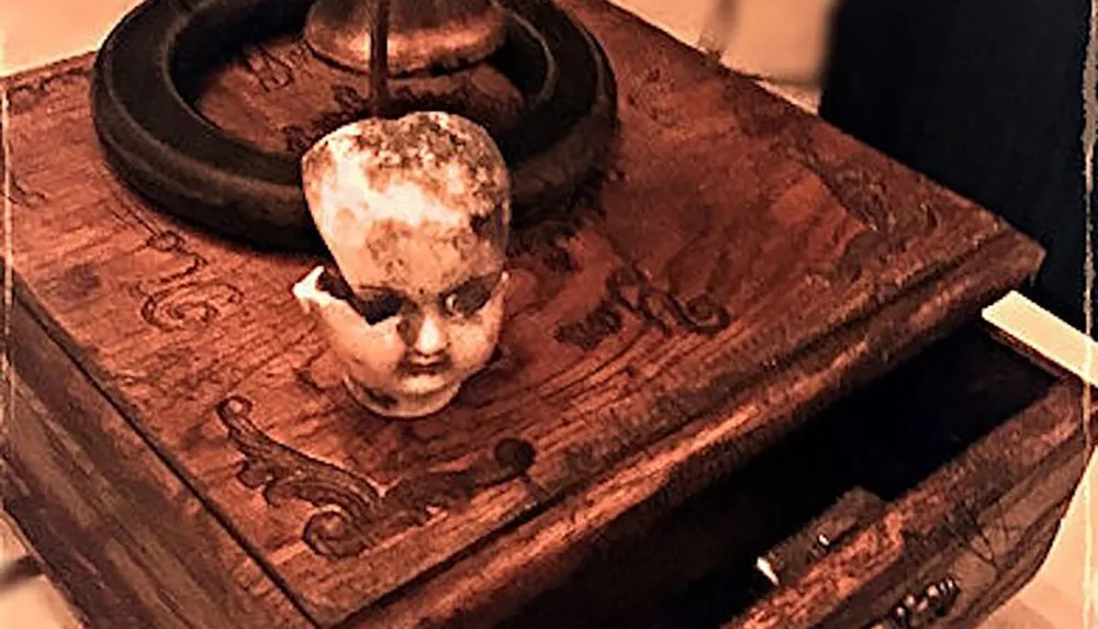 The image shows a weathered dolls head resting atop an old wooden box with intricate carvings creating an eerie atmosphere