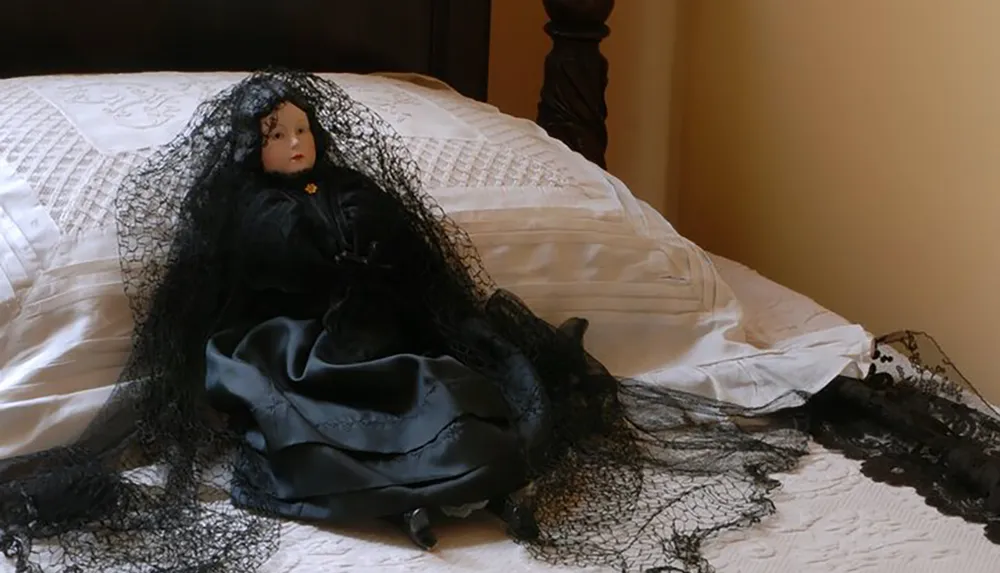 The image shows a porcelain doll dressed in black with a lace veil reclining on an all-white bedspread