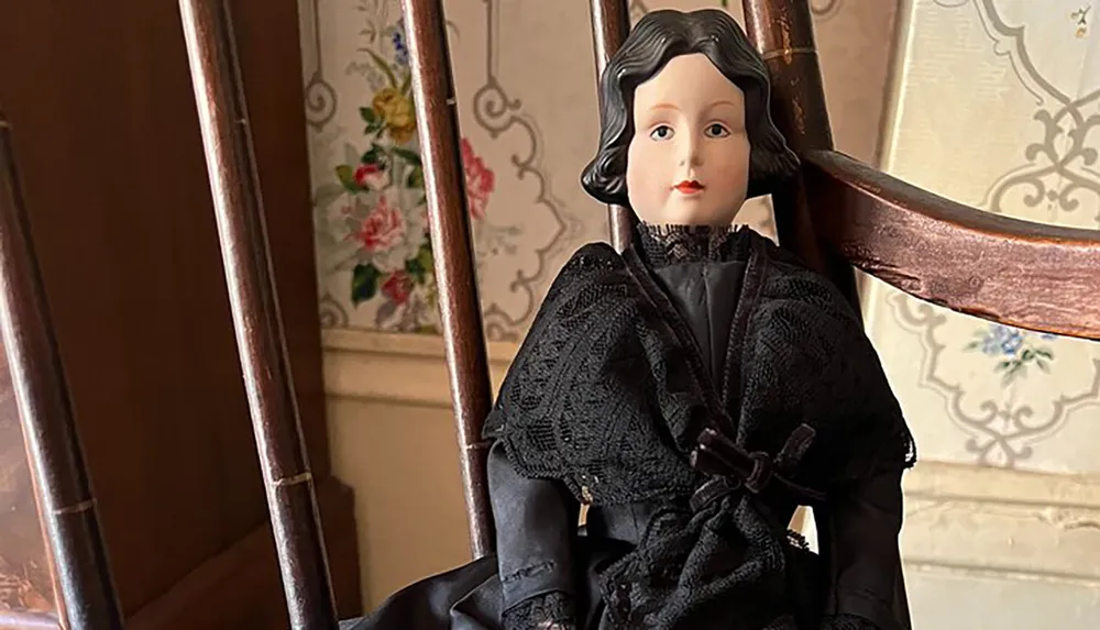 A porcelain doll with detailed facial features and wearing a black lace-trimmed dress is seated on a wooden rocking chair against a floral wallpaper background