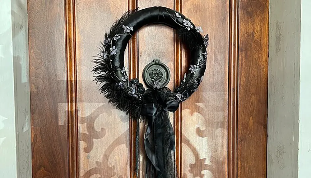 The image shows a decorative black wreath with feathers flowers and a bow hanging on a wooden door with ornate glass panels