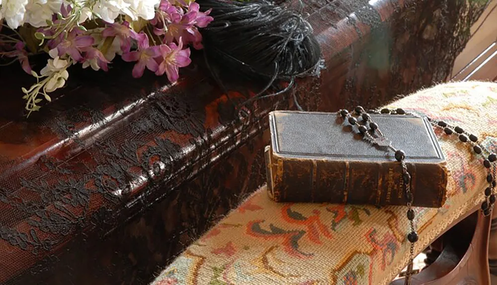 The image shows an antique book with a black beaded rosary on it resting on an embroidered cushion with a bouquet of white and purple flowers in the background suggesting a peaceful and historical ambiance