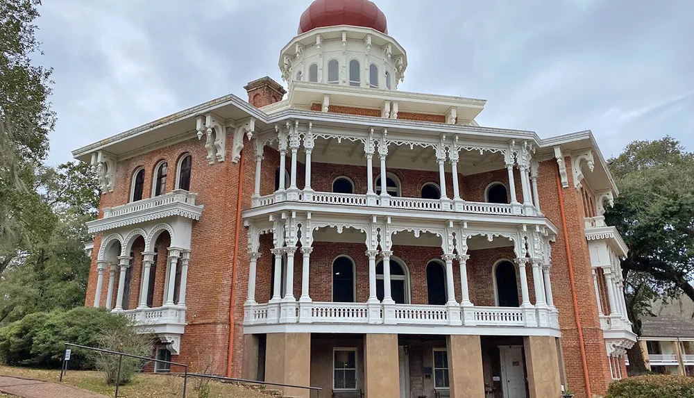 An ornate two-story brick building with white columns and balconies topped by a distinctive red dome appears stately against a cloudy sky