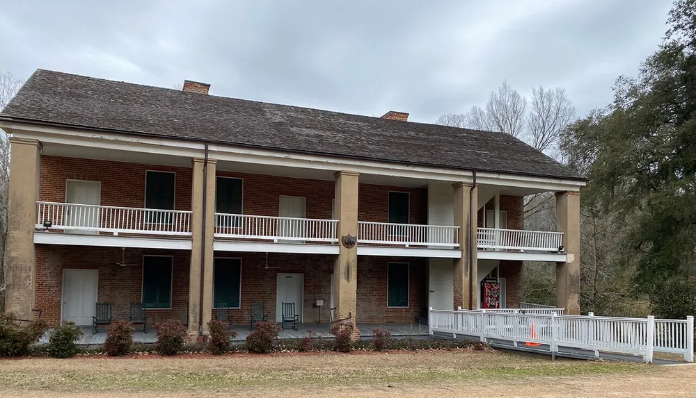 The image shows a two-story brick building with white columns and balconies surrounded by a white fence indicative of historic architecture