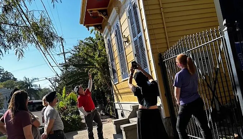 A group of people are gathered on a sidewalk with some looking up towards the corner of a yellow house while another person takes a photo