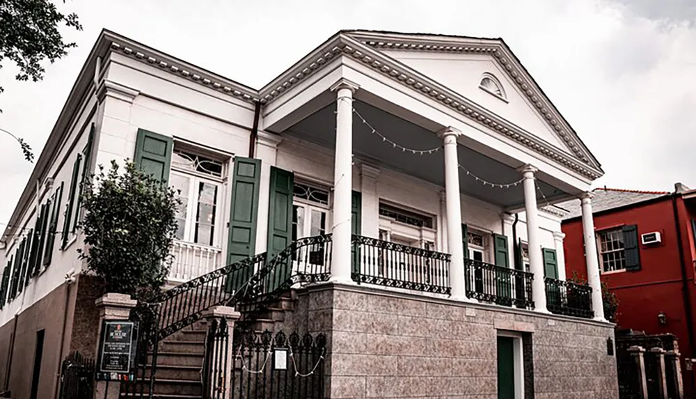 The image shows a classic two-story building with green shutters a balcony supported by columns and a staircase leading to the entrance situated next to a bright red building