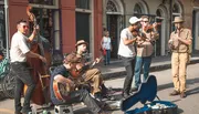 A group of musicians is performing live on a city street with instruments like a double bass, guitar, violin, and clarinet, as bystanders watch and a guitar case lies open to collect tips.