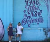 Two individuals are posing in front of a vibrant blue wall with the phrase One Time in New Orleans artistically painted on it