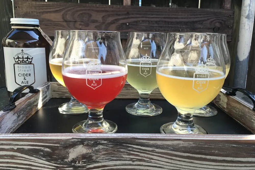 A flight of four different ciders is presented on a wooden surface each in a tulip glass with a branding that says Broad St Cider ALE with the first glass being a distinct red color compared to the others