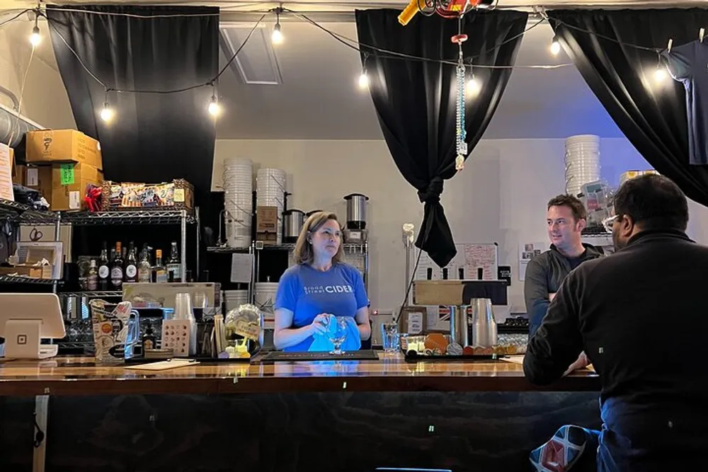 In this image a bartender stands behind the bar counter appearing to interact with two patrons seated on the other side