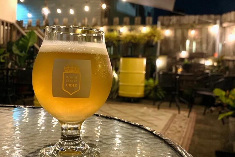 A glass of cider with a frothy head is prominently displayed on a table at an outdoor patio setting in the evening