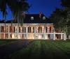 A two-story plantation-style house illuminated in the evening features a row of columns lining its front porches with Spanish moss hanging from nearby trees creating an atmospheric Southern scene