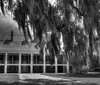 A two-story plantation-style house illuminated in the evening features a row of columns lining its front porches with Spanish moss hanging from nearby trees creating an atmospheric Southern scene