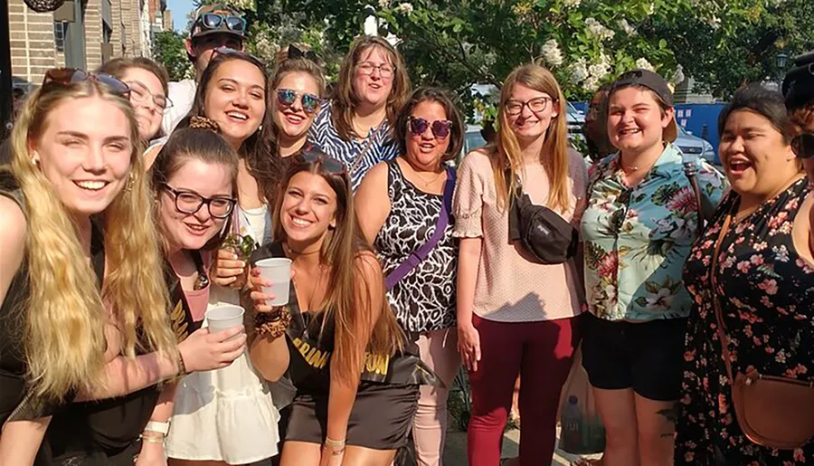 A group of cheerful people are posing together for a photo on a sunny day, some holding drinks and wearing sunglasses, suggesting a casual outdoor gathering or event.