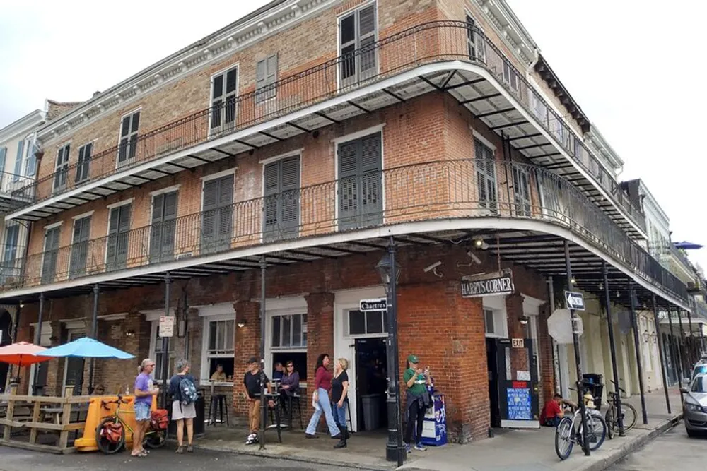 This image shows a group of people on a sidewalk by a traditional brick building with a wraparound balcony in an urban area possibly in a historical district like the French Quarter in New Orleans