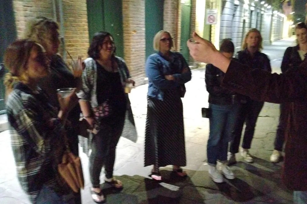 A group of people are standing on a street at night possibly engaged in a conversation or tour with one person pointing towards something of interest