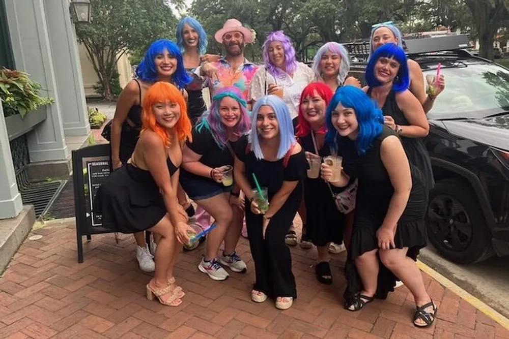 A group of people is posing for a photo on a sidewalk many with brightly colored wigs and drinks in their hands seemingly having a good time