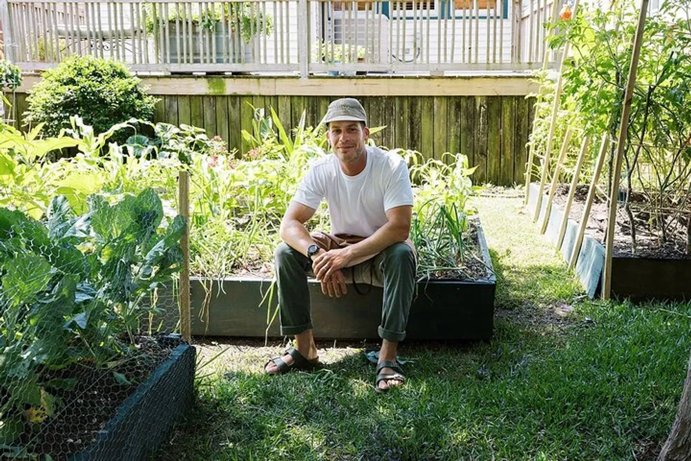 A person is sitting contentedly by raised garden beds filled with lush plants in a sunny backyard