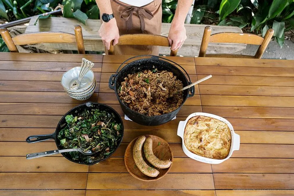 A person stands behind a wooden table set with a variety of dishes including what appears to be a pot of rice or grains a pan of greens a casserole and some sausages