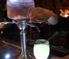 A persons hand is operating a spigot on a large glass beverage dispenser to pour a drink into a glowing cup in a dimly lit setting