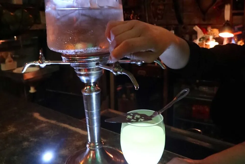 A persons hand is operating a spigot on a large glass beverage dispenser to pour a drink into a glowing cup in a dimly lit setting