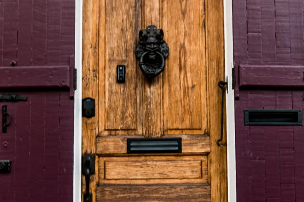 The image shows a close-up of a textured wooden door with a black lion-head door knocker in the center flanked by red shutters on either side