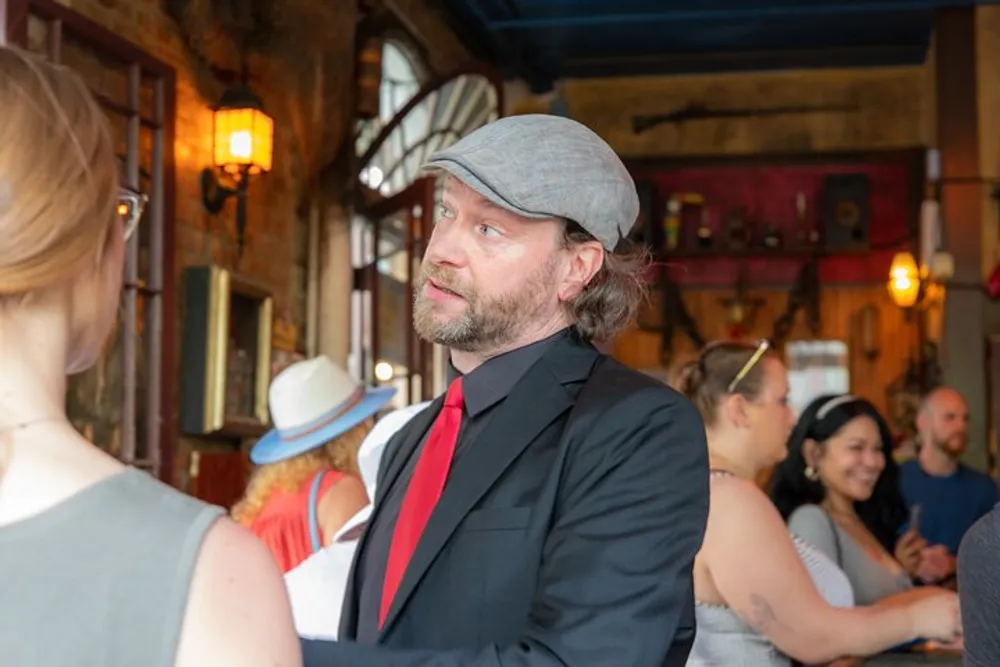 A man in a grey cap and black suit with a red tie is conversing with a woman in a social setting that appears to be a lively gathering