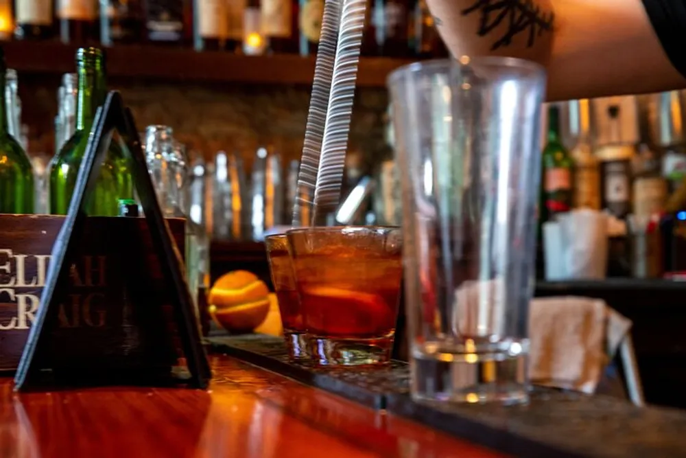 The image captures a close-up of a cocktail sitting on a bar counter with bottles in the background and a bartenders arm in motion suggesting a bustling bar environment